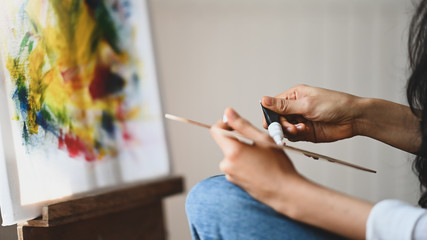 Cropped image of beautiful woman artist's hands while mixing color on the wooden color palette while sitting in front the painting canvas with modern art studio as background.