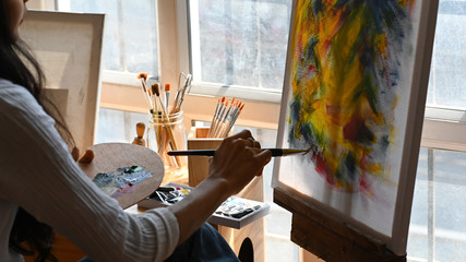 Cropped back image of young beautiful artist holding a brush and drawing on painting canvas while sitting next to the arts accessories putting on small wooden table with studio windows as background.