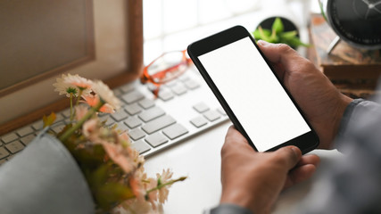 Plakat Cropped image of businessman's hands holding a cropped black smartphone with white blank screen at the modern working table with wireless keyboard, bouquet of flowers, eyeglasses, potted plant.