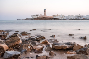 Fototapeta premium Sunrise on a rocky beach with a lighthouse in the background at Sur's bay, Oman
