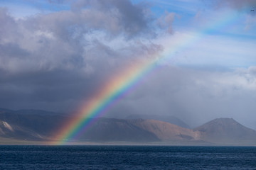 Beautiful summer landscape with sea, small village on the hill, bright colorful rainbow on blue sky, clouds and mountains on the horizon. Iceland, Peninsula
