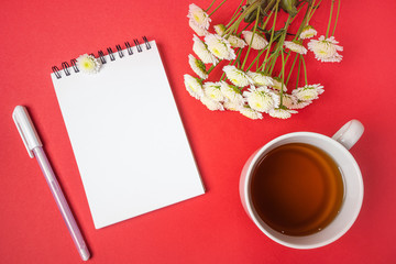 Notebook on a spiral with a blank page, pen, white small flowers and a cup of tea on a red background. Top view, flat lay, minimalism, copyspace.