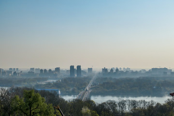 Naklejka premium A panoramic city view on Kiev from a hill. In the middle there is a Dnieper river and a bridge joining two shores of the river. The city is shrouded in fog.