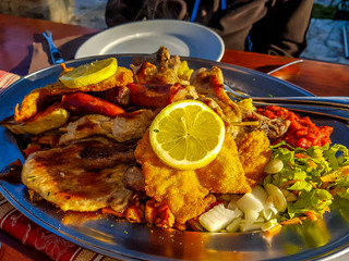 A silver plate filled with grilled chicken, fried fish and fresh salad, topped with fresh lemon. Sun shines bright on the meal, served in an open air restaurant.