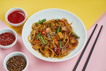 Appetizing homemade egg noodles in sweet and sour sauce with tofu and vegetables in a white bowl on colored background. Close up. Asian cuisine