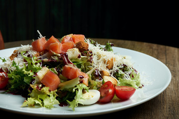 Close up view on served Caesar salad with smoked Salmon in white plate on wooden background. Tasty seafood salad, selective focus