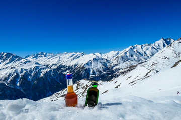Two bottles of liquor placed on a snow with high snow-capped mountains in the back. Apres ski in...