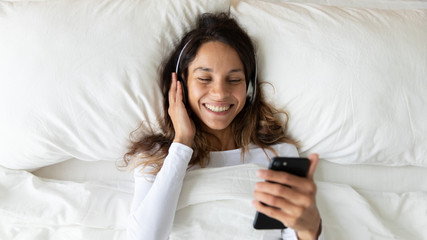 Top view of happy Caucasian millennial girl relaxing on white sheets in cozy bed listening to music on modern smartphone, smiling young woman wearing headphones have fun enjoy favorite tracks on cell
