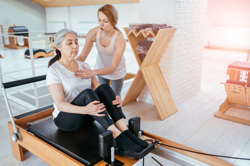 Brunette female instructor with cute two braids hairstyle consulting hansdome beard man workout pilates on reformer practice in pilates studio, working out indoor, correcting beginners, full length.
