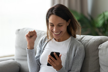 Excited millennial girl sit on couch in living room look at cellphone screen feeling overjoyed...