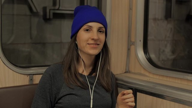Portrait of a cute girl in headphones and a blue hat on the subway, listening to music and browsing on a mobile phone in public transport. City lights background