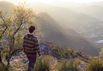 Side view of young man in casual clothes walking on grassy meadow at sunset. Beautiful landscape with mountains in the background.