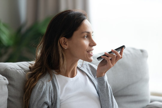 Young Caucasian Woman Sit On Couch In Living Room Using Smartphone Activate Virtual Digital Assistant, Millennial Girl Record Voice Message On Modern Cellphone Gadget, New Technology Concept