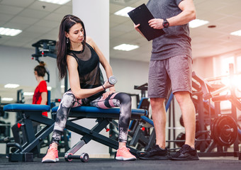 Sport, bodybuilding, lifestyle and people concept - young man and woman with barbell flexing muscles and making shoulder press squat in gym. Trainer and fitness girl are having workout at gym.