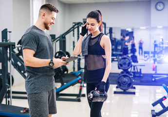 Fototapeta na wymiar Beautiful young woman with her personal trainer at the gym discuss her progress on a clipboard held by the man. Healthy lifestyle