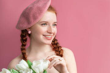Young happy smiling natural redhead lady with freckled skin, wearing pink beret, earrings, holding white tulips. Light pink color background. Spring beauty, fashion concept. Copy, empty space for text