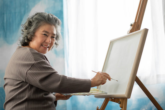 Happy smiling Asian Elderly woman oil painting on canvas at house on holidays.