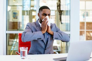 Portrait of a sick young man in business suit blowing his nose while sitting behind desk at work...