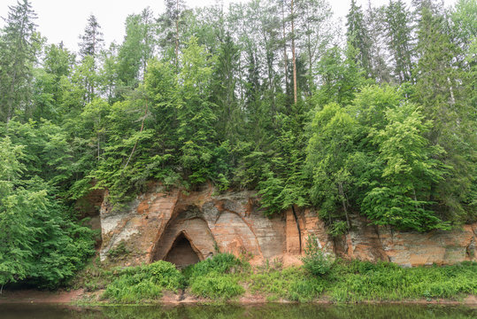 Sand stone cliffs and Salaca river. Red sandstones cave, water, trees and plants, green nature environment, climate concept..Angels cliff in Mazsalaca nature park, Latvia, Europe