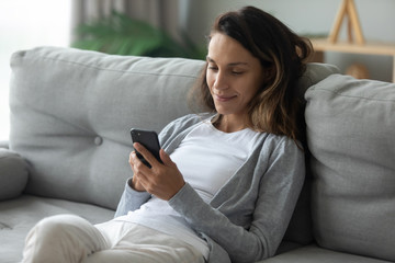 Smiling millennial Caucasian girl sit relax on cozy couch in living room browsing Internet on...