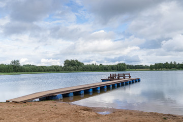 Fototapeta premium Wooden bridge for swimming on the shore of a lake. Blue sky and water, green trees, summer nature landscape. Environmental concept, Latvia, Europe.