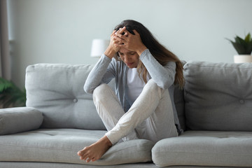 Fototapeta premium Depressed young woman it on couch in living room struggle with psychological trauma or mental illness at home, unhappy upset millennial girl crying struggle with depression after breakup or divorce