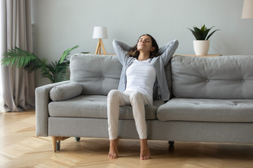 Happy millennial girl sit relax on comfortable couch in living room hands over head, peaceful young...
