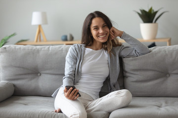 Obraz premium Portrait of smiling Caucasian millennial girl sit relax on comfortable couch in living room look at camera, happy young woman holding smartphone gadget rest on cozy sofa at home, technology concept