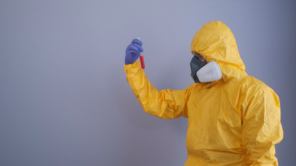 A man in yellow Hazmat suits and a respirator holds a test tube with a blood test, gray background.
