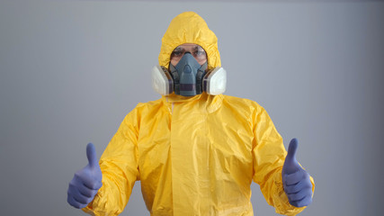 A man in yellow chemical protection suit and a respirator stands on a gray background and shows a thumbs up. - obrazy, fototapety, plakaty