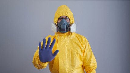 A man in yellow Hazmat suits and a respirator stands on a gray background with his arm outstretched and shows a stop sign with his hands.