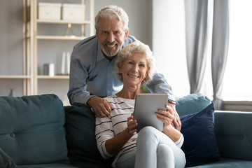 Portrait of smiling elderly 60s couple sit relax on cozy couch in living room using modern tablet together, happy old middle-aged 50s husband and wife look at camera browsing surfing internet on pad