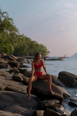 Sexy tanned brunette in red bikini sitting on the stone among tropical beach