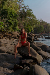 Fototapeta premium Sexy tanned brunette in red bikini sitting on the stone among tropical beach