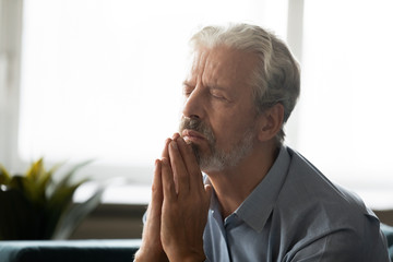 Calm hopeful elderly gray-haired man hold hands in prayer ask beg God for better future or fate, peaceful old 60s religious male believer Christian say worship pray at home, faith, religion concept