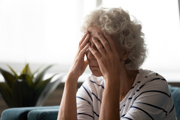 Upset old female hold head in hands feel distressed depressed suffering from solitude at home, unwell mature 50s woman touch temples having severe migraine or headache, elderly healthcare concept