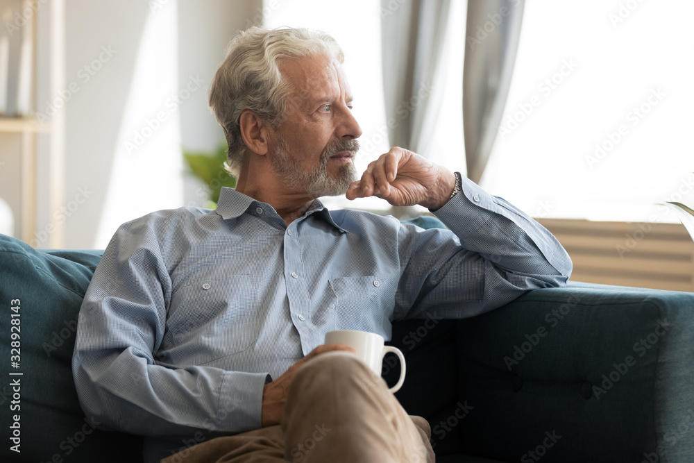 Wall mural pensive elderly 60s man sit relax on couch in living room drinking tea look in window distance think