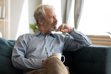 Pensive elderly 60s man sit relax on couch in living room drinking tea look in window distance...