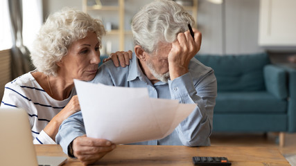 Distressed mature 60s husband and wife sit at table calculate manage household expenses having...