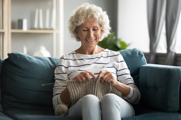 Happy mature 60s grandmother sit relax on couch in living room knit at home, smiling middle-aged...