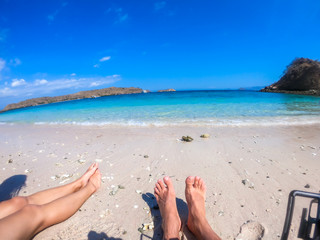 Fototapeta na wymiar Two pairs of legs resting on an idyllic white sand beach on Komodo Islands, Indonesia. The beach is gently washed by the waves. There are some cliffs on the side. Relaxation and chilling