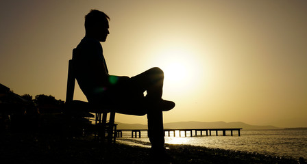 Obraz premium Young sad depressed man sit alone on chair, looking at distant sea / seascape horizon. Time to go, say goodbye. Miss someone. Desire, hope to go far away. Unhappy boy feel lonely at seaside outdoor. 
