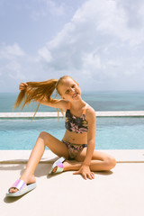 A small and beautiful girl with red hair and freckles sits by the pool in a bathing suit in the villa and poses in front of the camera. Children's fashion
