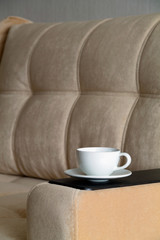 white cup of coffee on the arm of an armchair. details of brown sofa in the living room. take a break