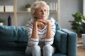Thoughtful old 60s lady sit on couch in living room look in distance remembering missing past,...