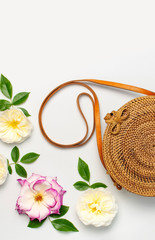 Fashionable concept of women's handbags. Handmade natural organic rattan bag and delicate rose flowers with green leaves on light background. Flat lay, Copy space, top view. Ecobags from Bali