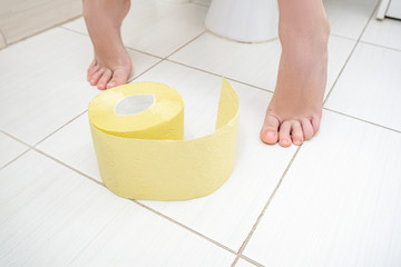 Cropped legs of a boy sitting on a toilet with a roll of yellow toilet paper on a floor, digestive problems and difficulties in toilet concept