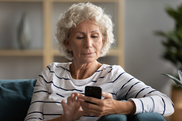 Modern mature 50s woman sit on couch in living room texting messaging on smartphone, smart...