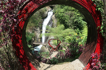 A red heart-shaped frame seen through red round frame with Gitgit Twin Waterfall in the back, Bali Indonesia. Touristic attraction for taking selfies with the waterfall. Lush green flora around.