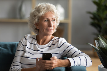 Happy old 50s woman sit on couch at home using modern cellphone gadget look in distance thinking,...